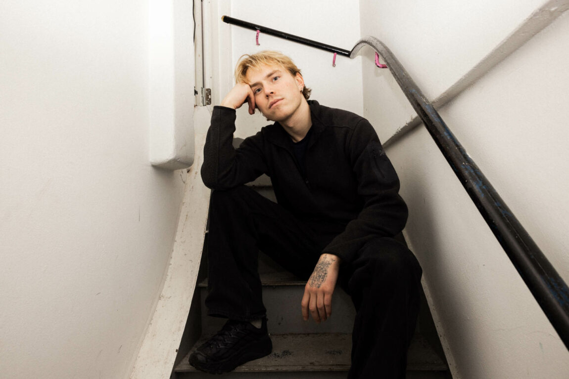 Artisten Fig Tape sitter i en smal trapp iført helt svarte klær. Han lener siden av hodet sitt på knyttneven sin.