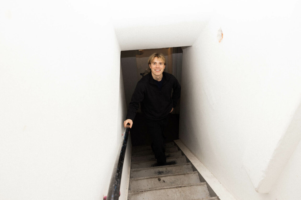 Artisten Fig Tape står nederst i en smal trapp og smiler bredt. Han har på seg en svart genser og bukse.