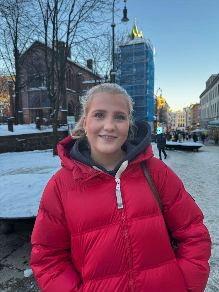 Karoline Thorsen ser smilende inn i kamera. Hun står på Karl Johan i Oslo med mennesker vagt i bakgrunnen. Et av menneskene bak henne er anonymisert med NPC meme ansikt som er tegnet over hans ansikt. Memen har små prikker til øyne, trekantnese og strekmunn.