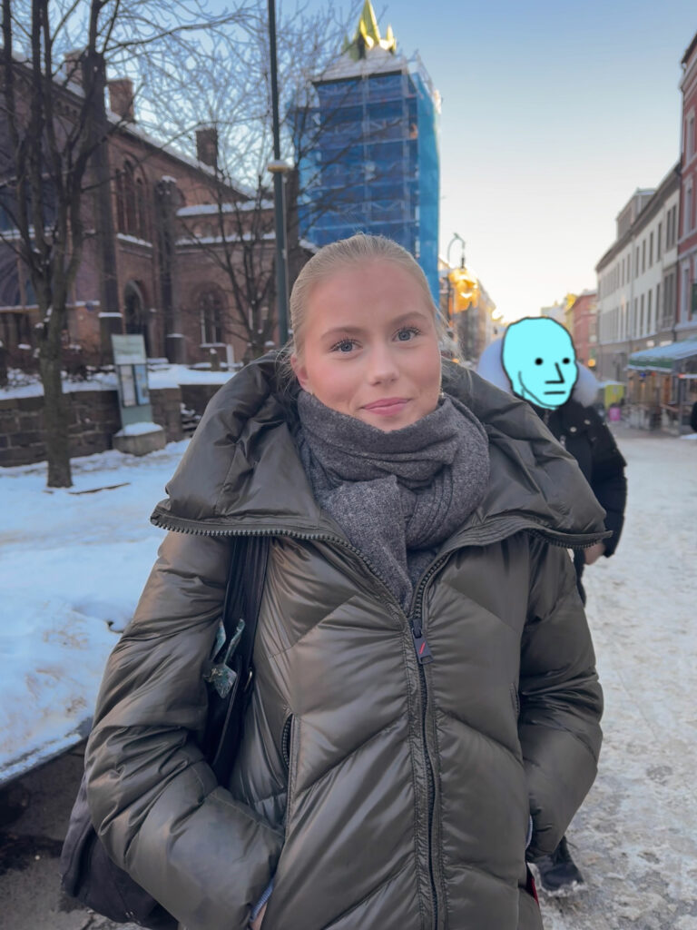 Maren Ohlgren ser smilende inn i kamera. Hun står på Karl Johan i Oslo med mennesker vagt i bakgrunnen. Et av menneskene bak henne er anonymisert med NPC meme ansikt som er tegnet over hans ansikt. Memen har små prikker til øyne, trekantnese og strekmunn.