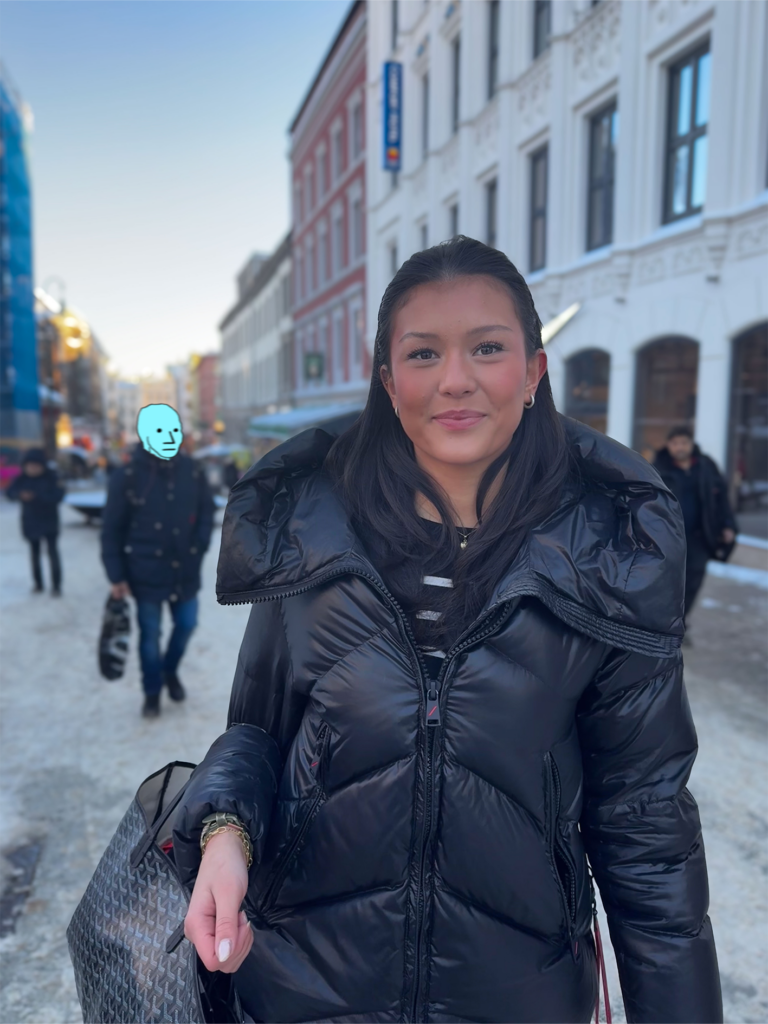 Caroline Skovli ser smilende inn i kamera. Hun står på Karl Johan i Oslo med mennesker vagt i bakgrunnen. Et av menneskene bak henne er anonymisert med NPC meme ansikt som er tegnet over hans ansikt. Memen har små prikker til øyne, trekantnese og strekmunn.