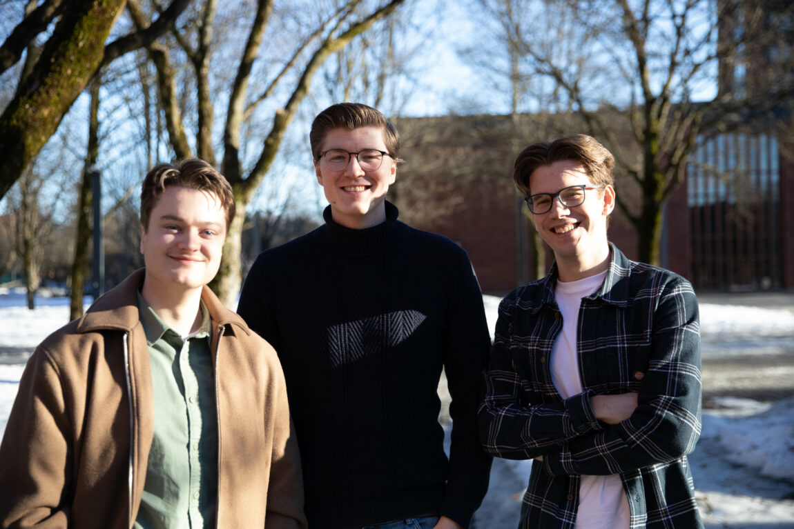 Bilde av tre mannlige studenter ved Universitetet i Oslo. De står skulder til skulder og smiler til kamera. Han korteste av dem til venstre har på seg grønn skjorte og brun åpen jakke. Han  i midten har på seg en mørk turtleneck og briller. Han til høyre har på seg en åpen og rutete skjorte og har også briller.