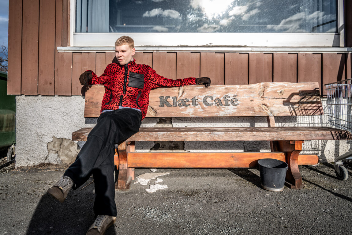 Ung mann som sitter på en benk i sola. På benken står det "Klæt Cafe". Han har på seg en rod fleece med leopardmønster.