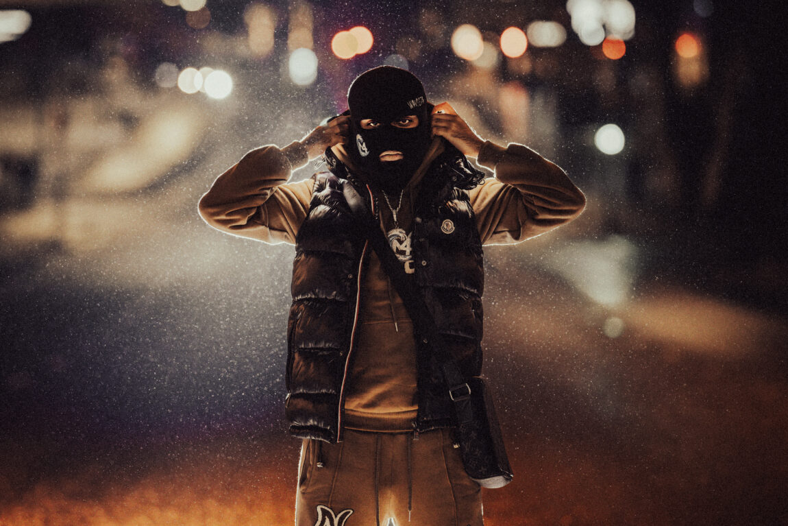 Bilde av rapperen M4 som står ute i regnet med en svart finlandshette og retter på hetta si. Han ser rett inn i kamera med et morskt blikk.