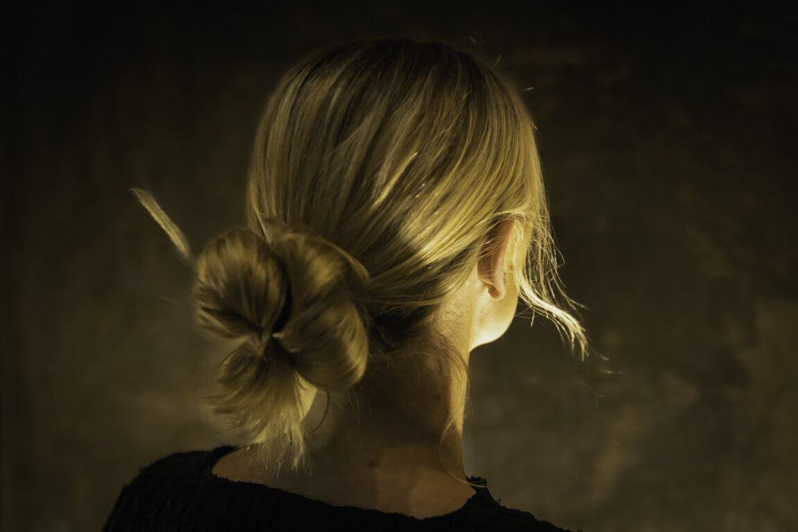 Bildet viser "Synne" sett bakfra. Hun har blondt hår som er satt opp i en knute. Bildet er ganske mørkt, og vi ser ikke ansiktet hennes. 