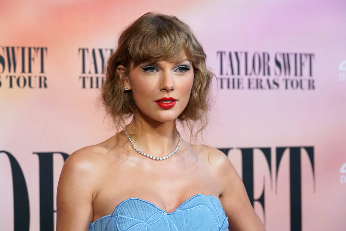 Et bilde av Taylor Swift foran en rosafarget filmplakat. Hun ser halvveis smilende, halvveis alvorlig ut. Hun har på en blå kjole eller topp.