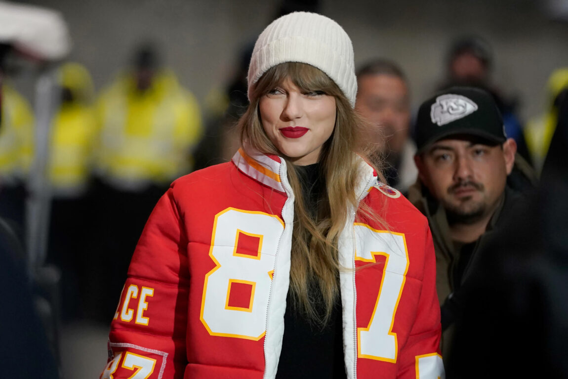 Taylor Swift har på seg en rød jakke og hvit lue. Hun smiler lurt og ser til siden.
