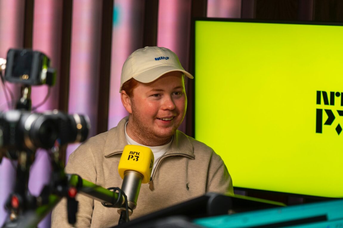 Snorre sit i eit P3-studio med ein stor gul skjerm bak seg. Det er ein gul P3-mikrofon retta mot han. Snorre har oransje hår, beige caps med blå skrift på og ein beige genser med kvit t-skjorte under. Han smiler mot høgre for kamera. 