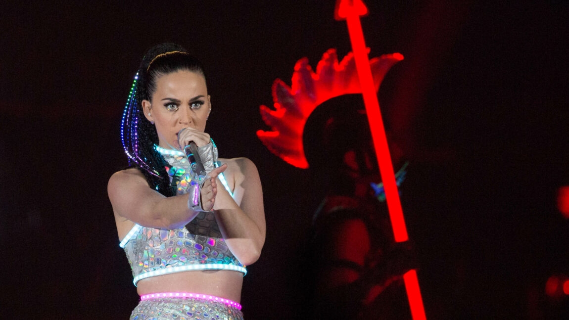 Katy Perry er på scenen med en mikrofon og en glitrende kjole med lysende neondetaljer. Hun peker på publikum.