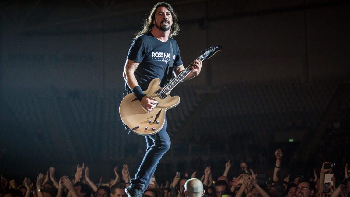 Gitaristen i Foo Fighters står på en scene og spiller gitar mens han går fremover. Har på seg en svart t-skjorte og har langt svart hår.