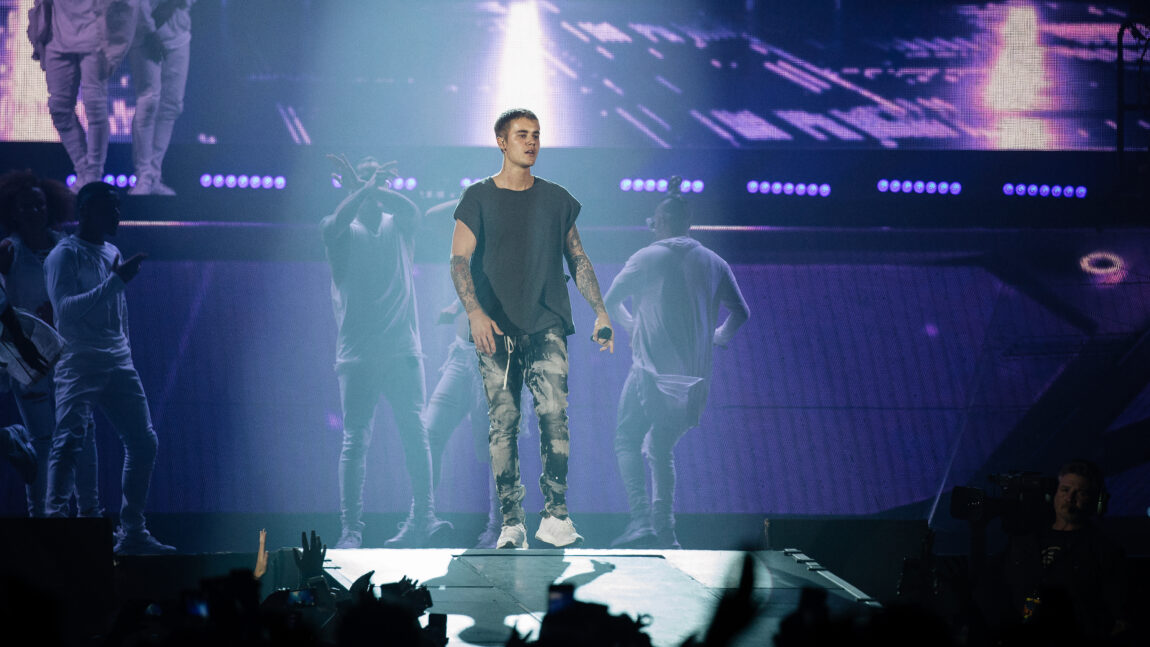 Justin Bieber har på seg en svart t-skjorte og står på scenen med en mikrofon i hånde, mens han ser på publikum. 