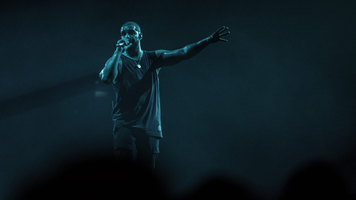 Drake i en svart singlet. Holder en mikrofon i høyyre hånde og holder venstrehånden ut mot publikum. 