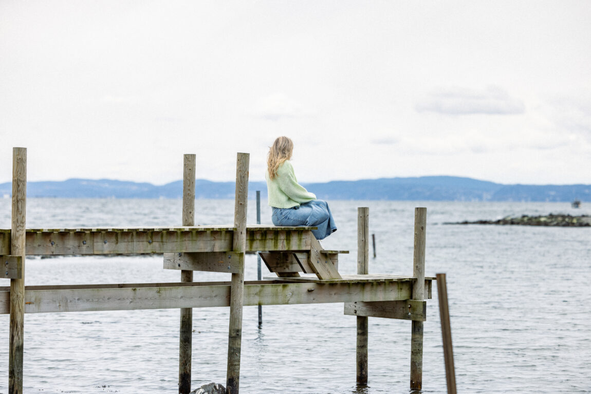 Maud sitter på en brygge av tre og ser ut mot sjøen. Vi ser bare bakhodet hennes bakfra.
