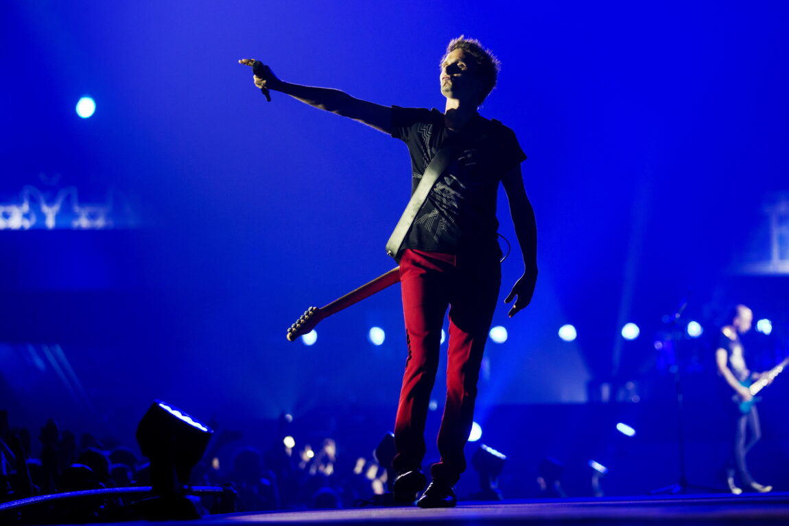 Gitaristen i Muse står på en scene og peker mot publikum, mens han lener seg bakover.