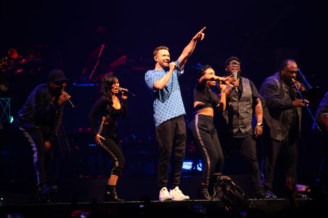 Justin Timerlake står på scenen med en blå t-skjorte og peker mot publikum mens han synger. Ved siden av han står det flere korister.