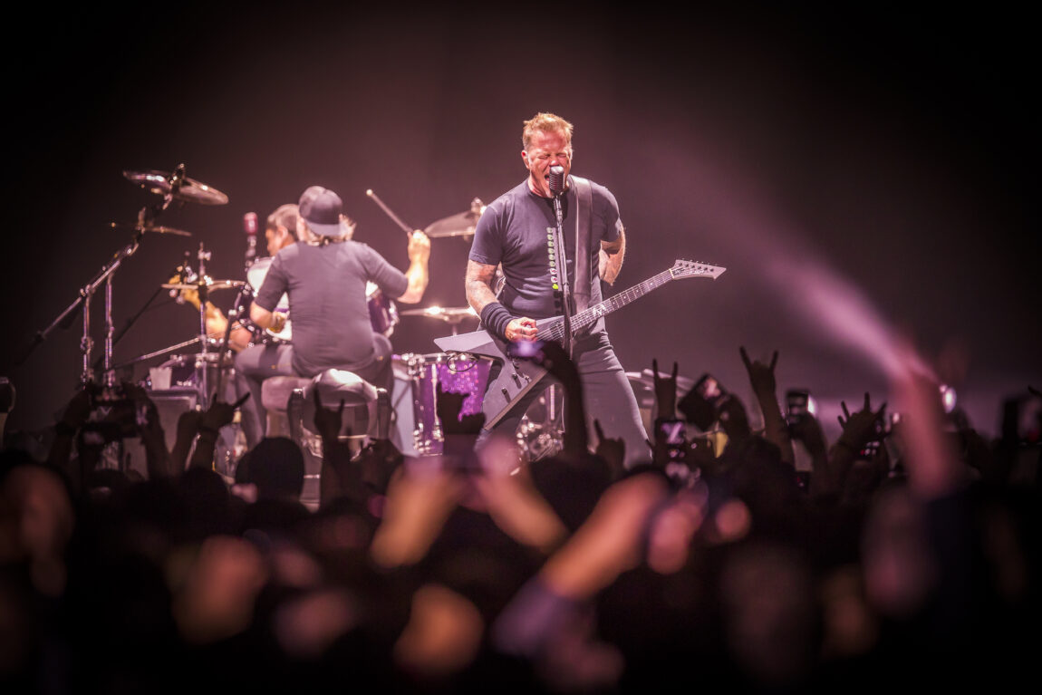 Gitaristen i Metallica synger intenst inn i mikrofonen. Vi ser publikum hender i bakgrunnen. Bak gitaristen spiller trommeslageren med ryggen til.