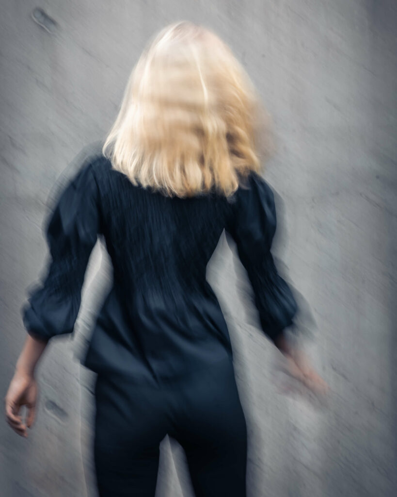 Bildet viser "Synne" sett bakfra. Hun har blondt hår som henger løst, og har på seg svarte klær. Bildet er ganske blurry og tatt i bevegelse. 