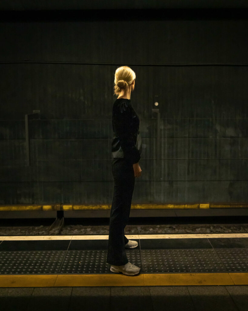 Bildet viser "Synne" sett bakfra. Hun har blondt hår som er satt opp i en knute. Hun står ved siden av et t-banespor. Hun har på seg svart bluse, svart bukse og hvite joggesko. 