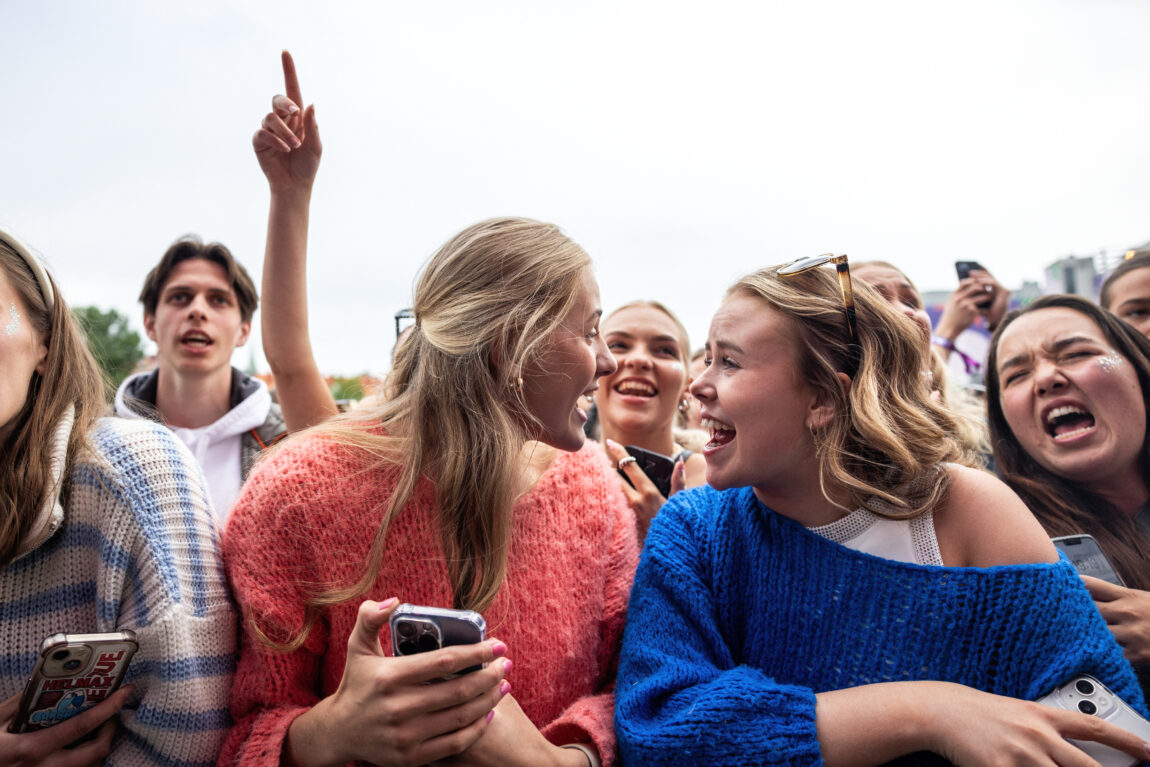 To kvinnelige fans står på første rad og smiler mot hverandre. Hun til venstre har på seg en oransje/rosa genser og hun til høyre har på seg en mørkeblå genser. I bakgrunnen ser man andre fans som ser tydelig engasjerte ut.