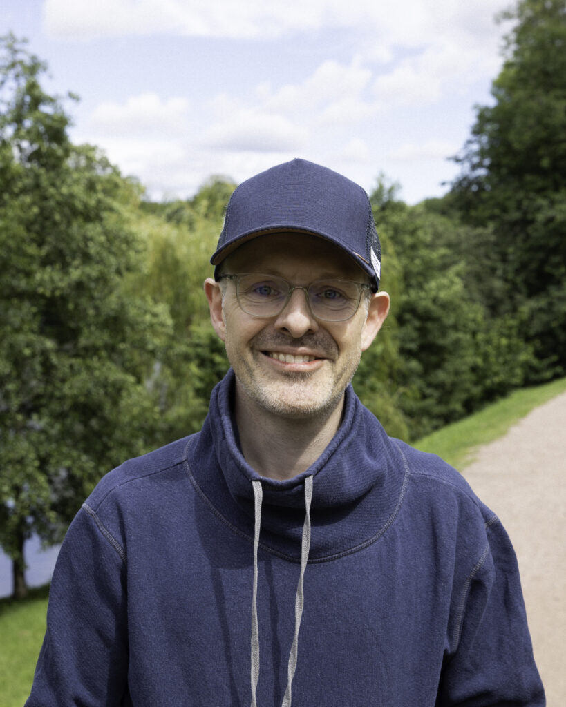 Middelaldrende mann med blå hettegenser, caps og briller. Han står og ser inn i kameraet med et lurt smil i en park. 