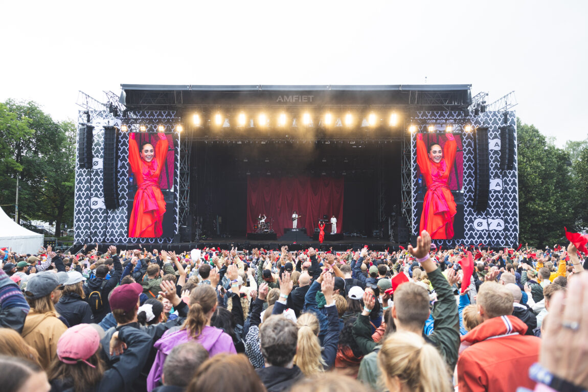 Oversiktsbilde av scene og publikum. En kvinne kledd i rødt kan ses på skjermene på scenen. Flere har på seg regnjakke i publikum.