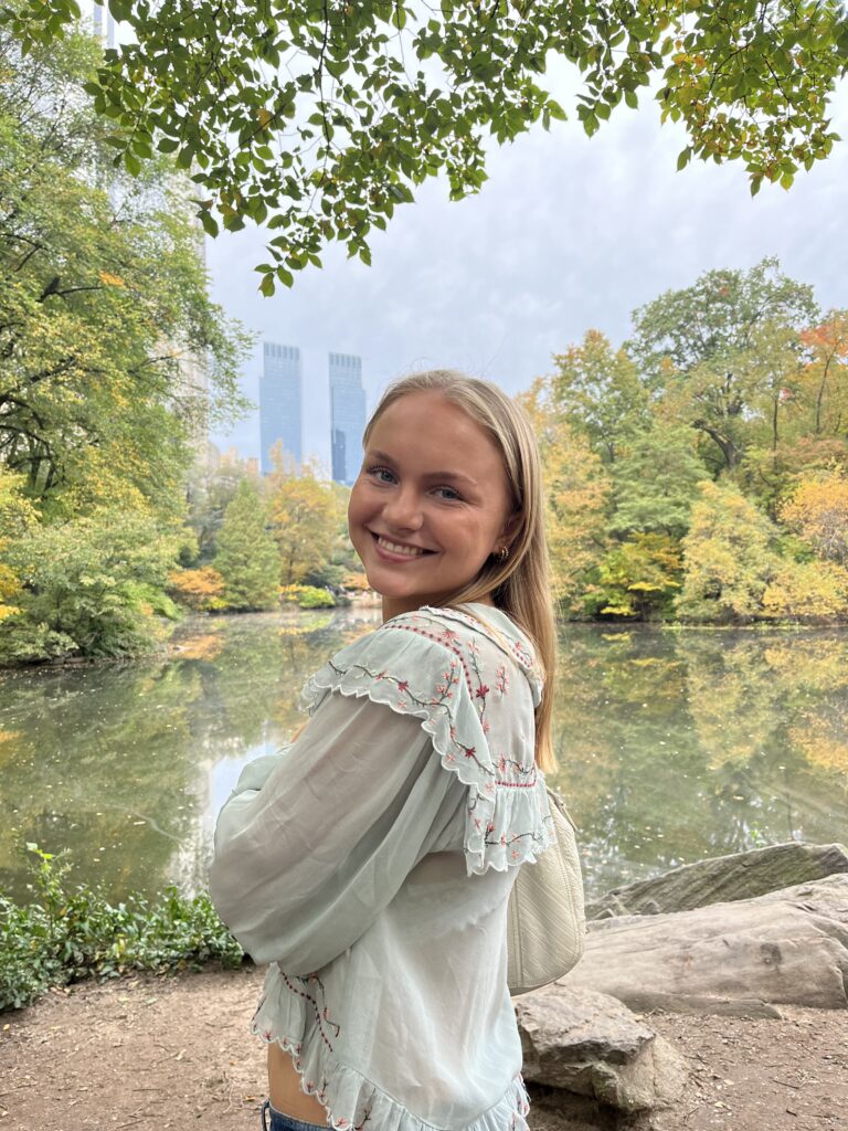 Selma Løvendahl står med langt blondt hår og ser seg bak over skuldren inn i kamera. Bakgrunnen er Central Park i New York. Selma står foran et vann og fargene er grønne og skiftende til gule høstfarger på trærne.