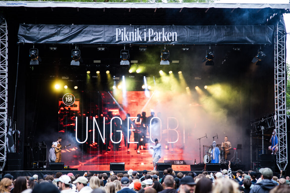 Bakgrunnsbilde av en Unge Obi konsert. Ved siden av han står det en bassist og en trommis. De har på seg oransje tracksuites. Obi har på seg en blå skjorte og shorts og en caps.