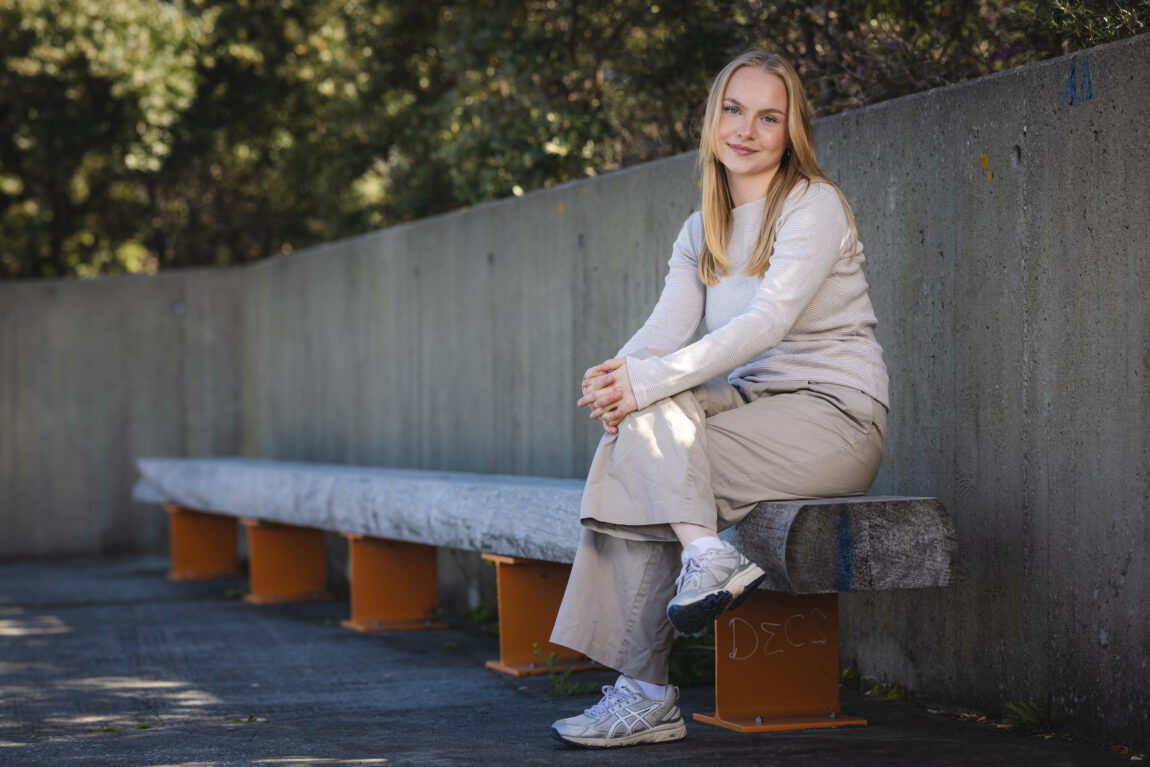 Blond jente sitter med beina i kryss på benk. Betongvegg i bakgrunn. Hun har langt blondt hår og smiler med lukket munn. Solen skinner, hun sitter i skyggen.