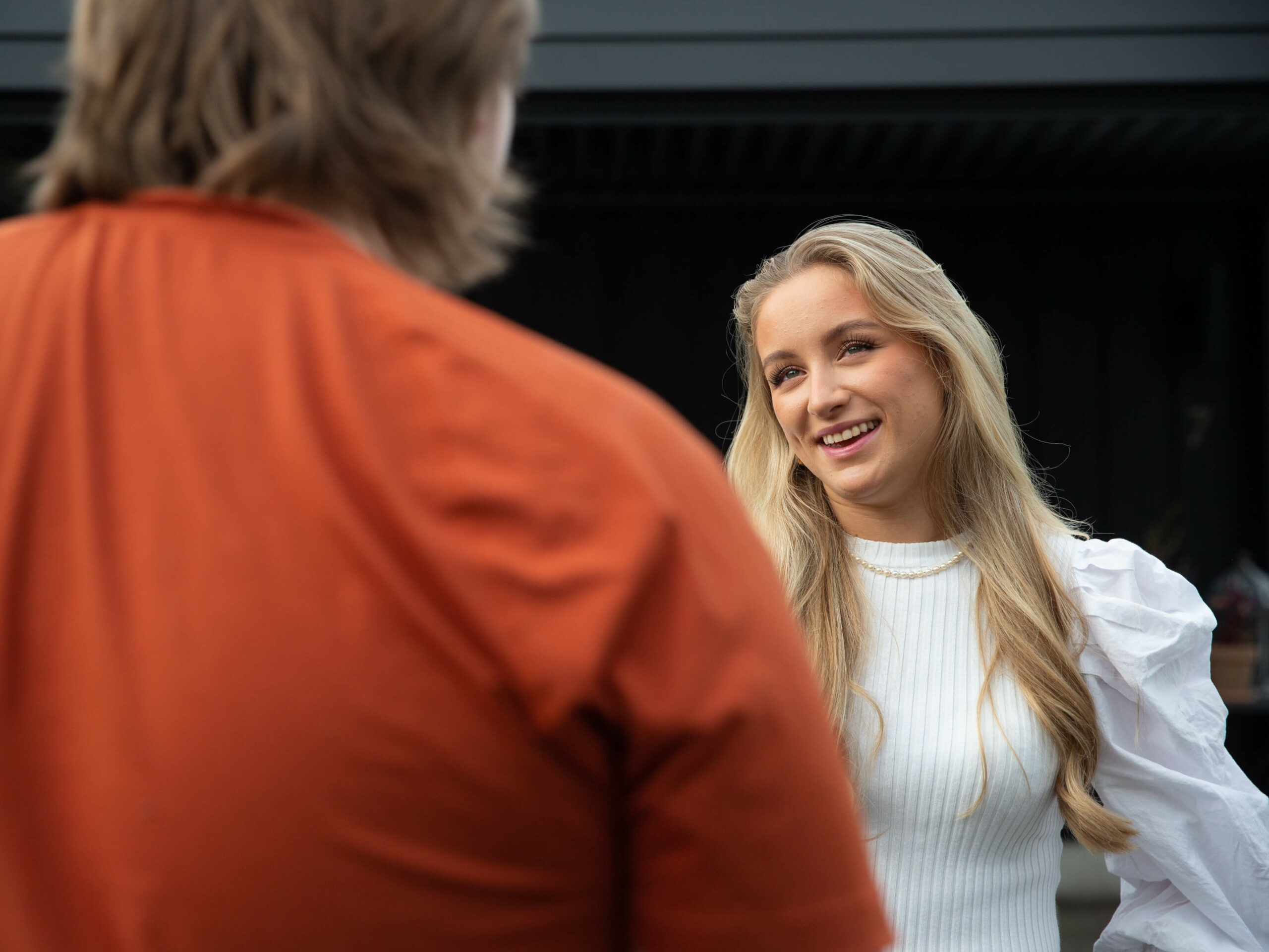 Bildet viser Maria, en ung kvinne med langt lyst hår og hvit bluse. Hun snakker med en annen person, som vi bare ser ryggen til. Denne personen har på seg rød genser.