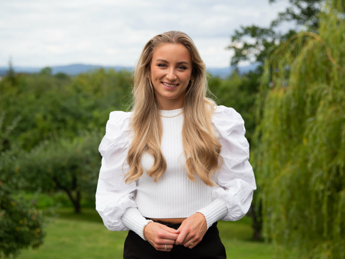 Biletet viser Maria Kleven. Ho har langt lyst hår, kvit bluse og svart bukse. Ho ser rett i kameraet og smiler. I bakgrunnen ser ein grøne tre.