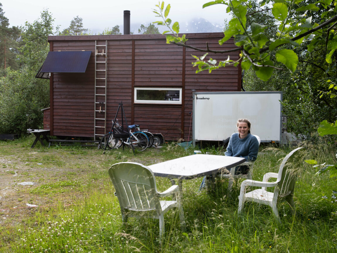 Anna sit framfor minihuset sitt i ein plaststol. Det er to andre plaststolar og eit stort bord. Minihuset er brunt, har ein stige opp langs eine veggen, solcellepanel og ein stor kvit hengar framfor seg. Det er grønt i graset rundt og skyer dekker fjellet bak ho. 