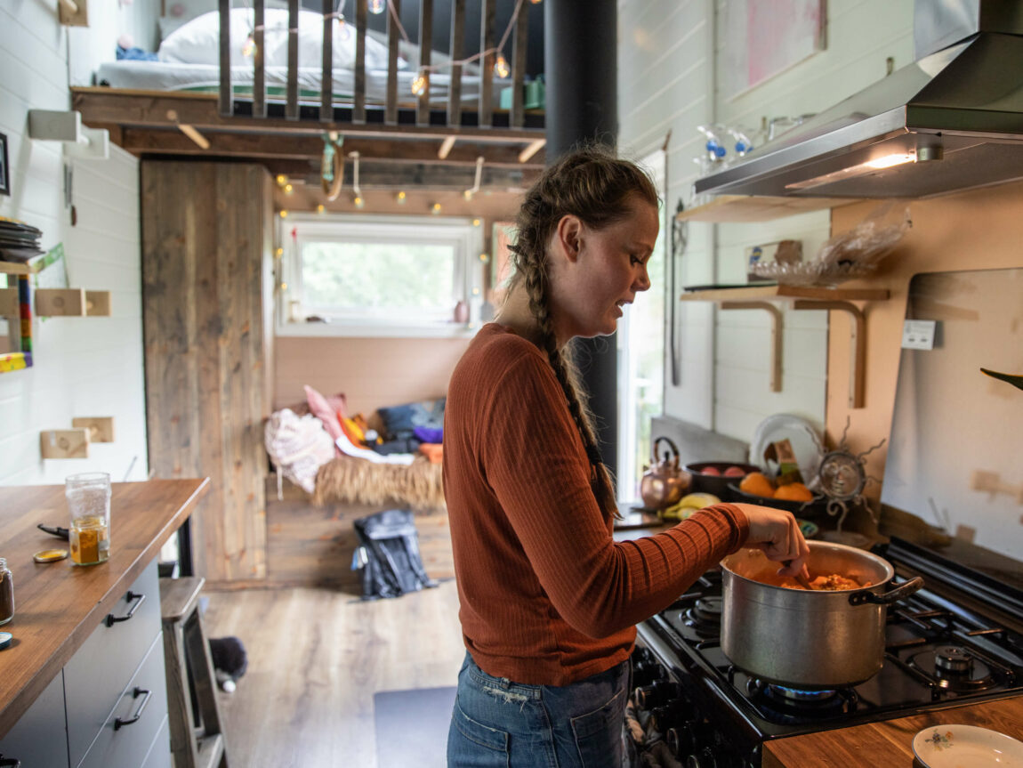 Anna står og rører i gryta med mat mens ho snakkar. Bak ein ser ein andre halvdelen av minihuset, som består av ein benk og ien hems.