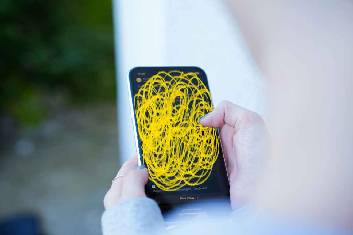 Viva holder i telefonen sin. Hun har søkt på "magesmerter", men store deler av telefonen har en gul krusedull over seg. Bildet er tatt fra bak skulderen hennes.