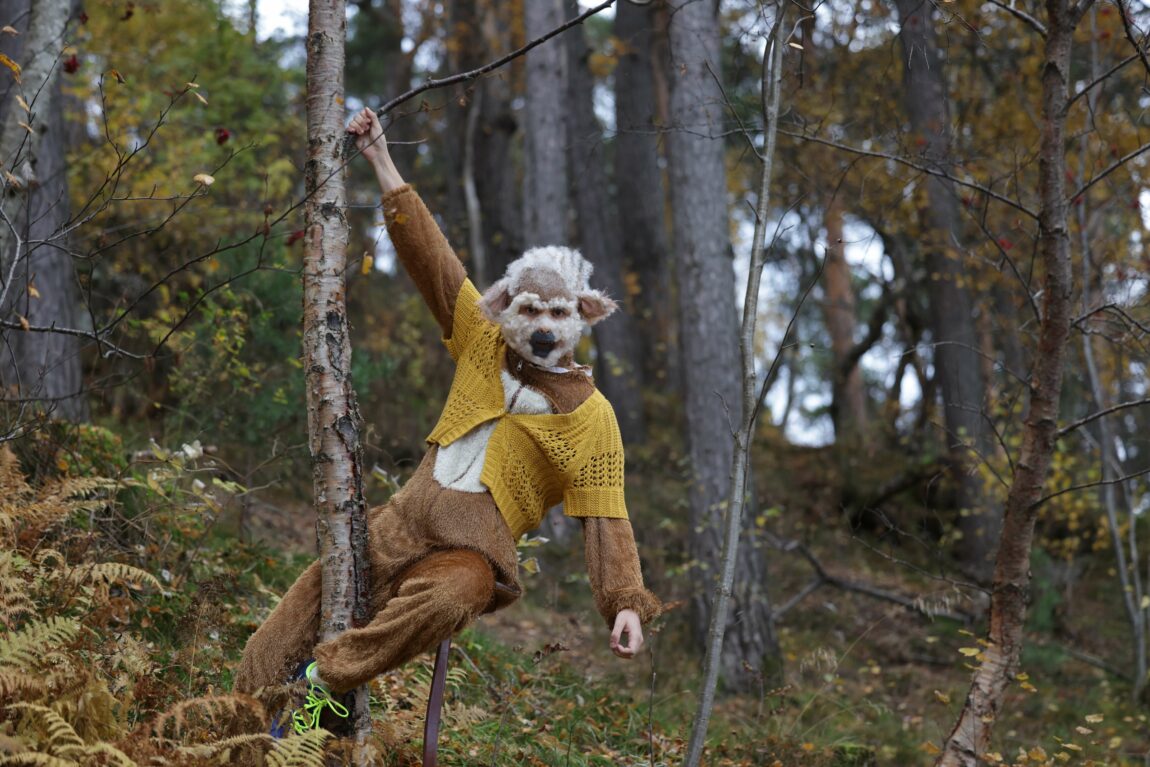 Henning henger i et tre med en arm. Han er sminket som en ape. I fjeset har han det som skal forestille ape-hår. Han ser rett i kameraet.