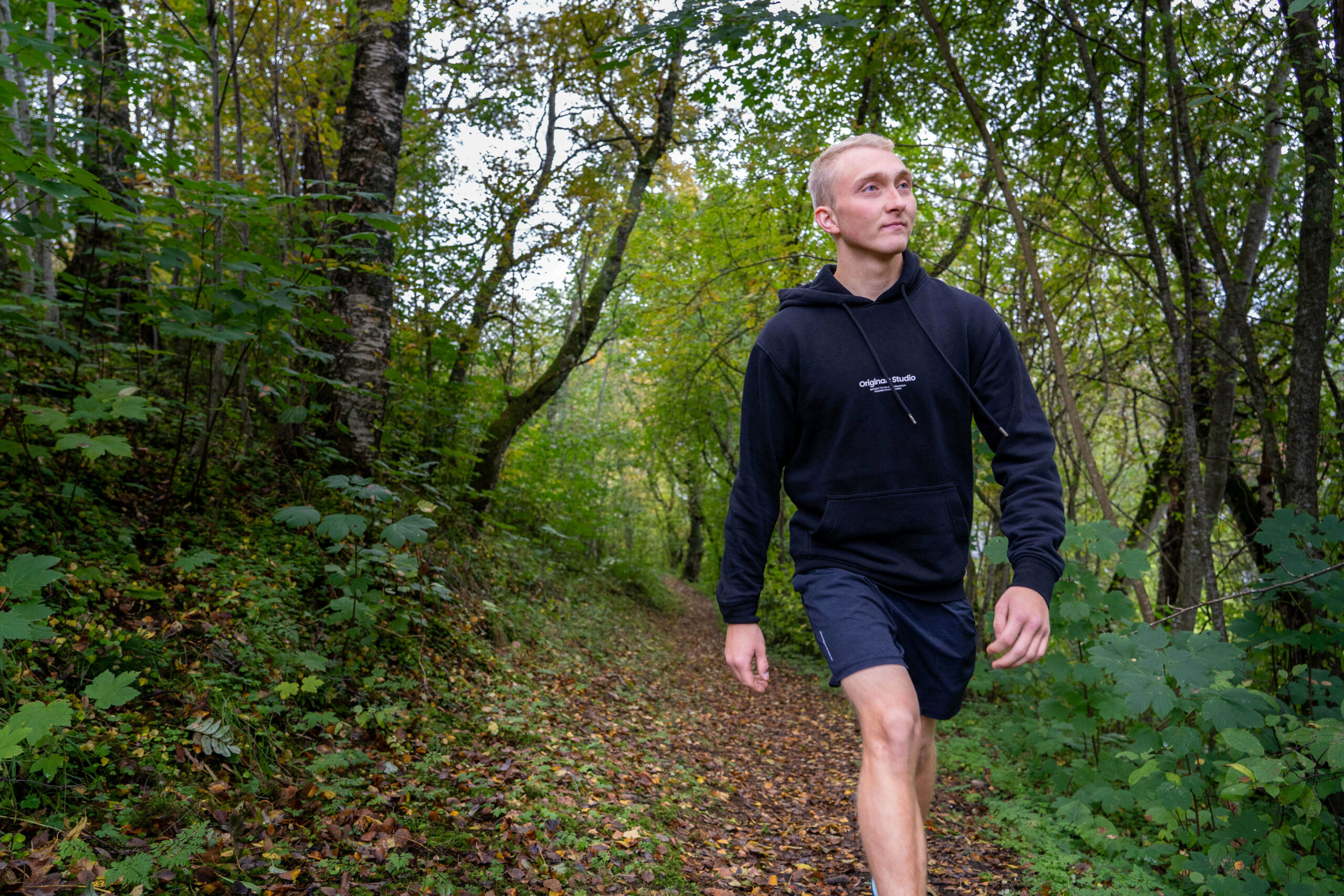 Bildet viser en ung mann, Michael, som går langs en skogssti omgitt av høstfargede trær og løv. Personen har blondt kort hår, og han har på seg en svart hettegenser og mørke shorts, og er iført joggesko. Skogsstien er dekket av falne blader, og det er tidlig høst. FOTO: Jenny Westrum-Rein, NRK P3