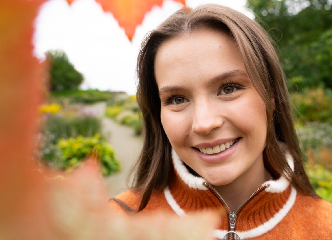 Nærbilde av Emma som ser smilende i kameraet. Foran henne er det et oransje løv.