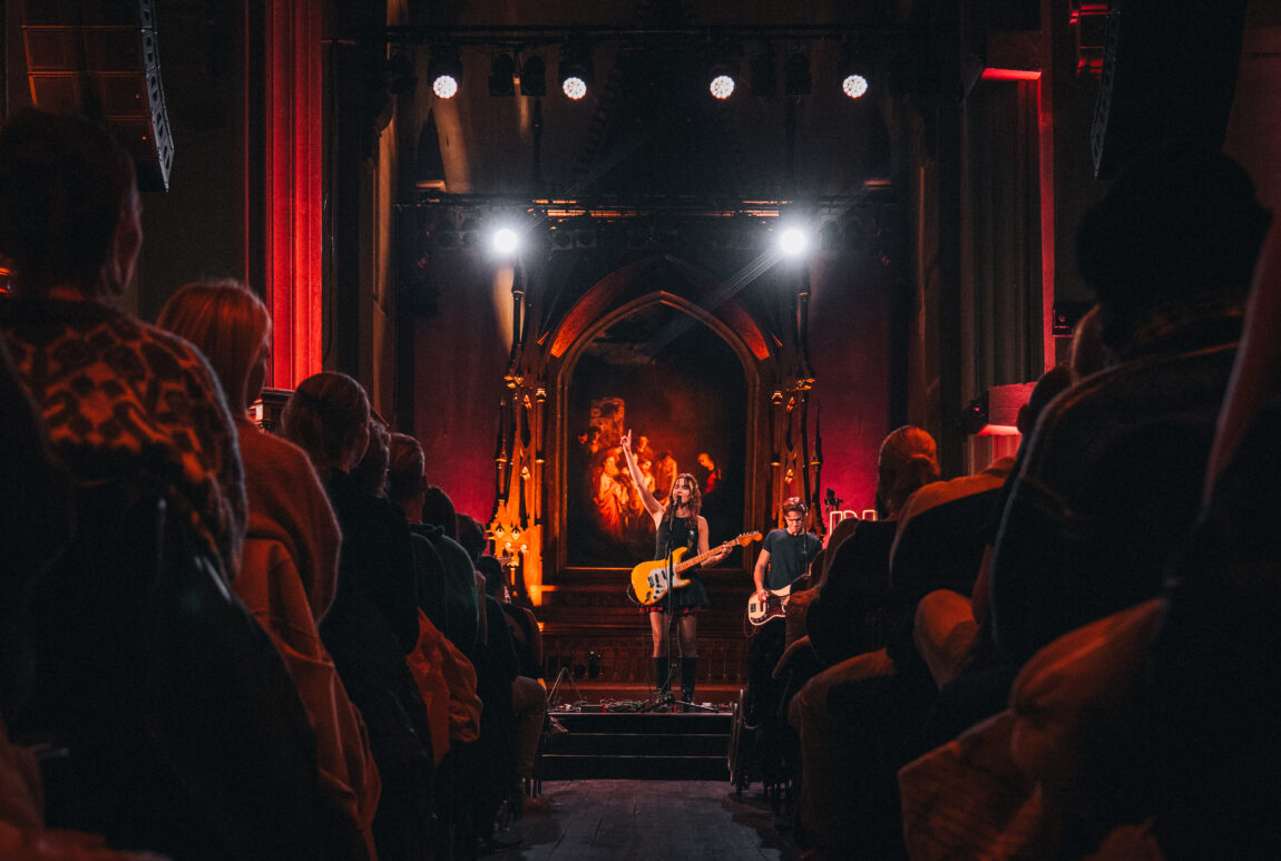 Anna Lille står på en scene og spiller gitar og peker i været. Gitaren er grønn. Vi ser bakhodene til flere i publikum. (Som av en eller annen grunn har fått sitteplasser??)