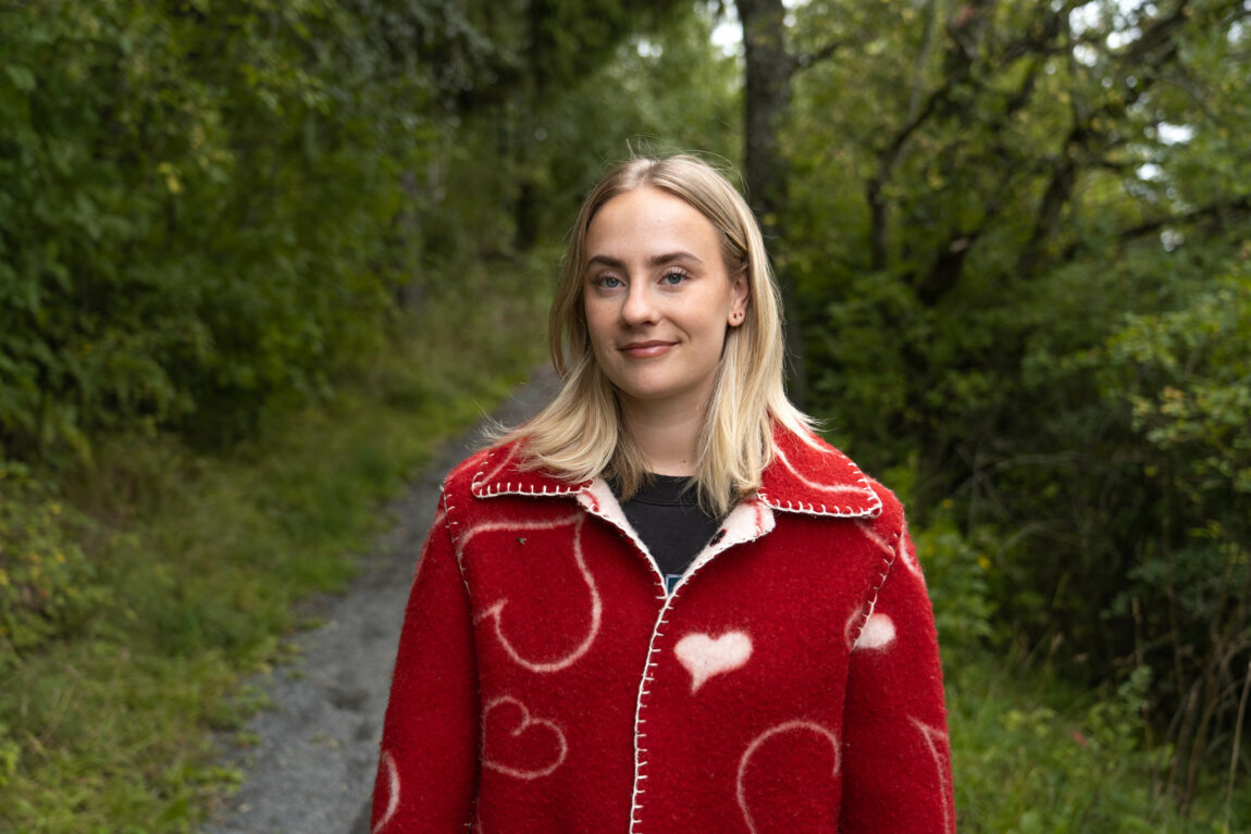 Oda ser i kameraet og smiler. Hun har blondt hår som går ned til skuldrene. Hun har på seg en rød ull-jakke med hvite hjerter på.