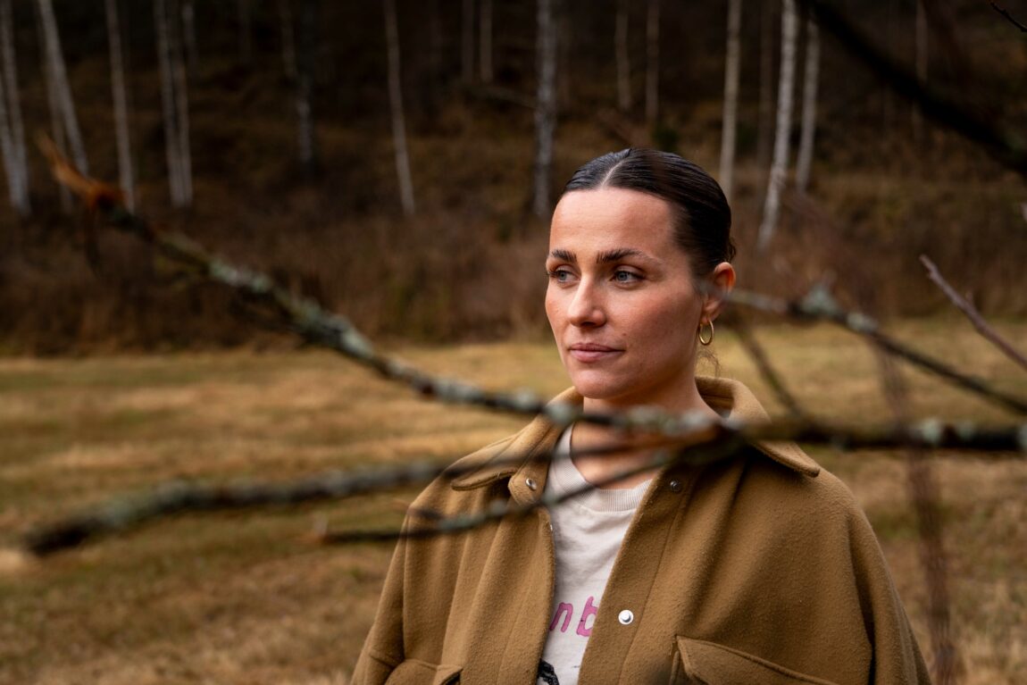 Bildet viser Karoline, en ung kvinne med mørkt hår i en lav hestehale. Hun har på beige jakke,  hvit skjorte og store øreringer i gull. Foran henne ser man noen nakne greiner. Hun ser til venstre for seg med et alvorlig uttrykk. 