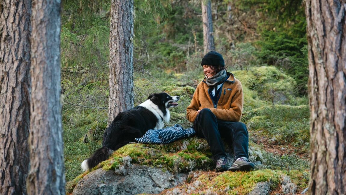 Fay Wildhagen og den svarte og hvite hunden hennes løper rundt henne. De sitter mellom skog og gress ute i naturen. Fay har på seg en oransje jakke-genser og svart lue.