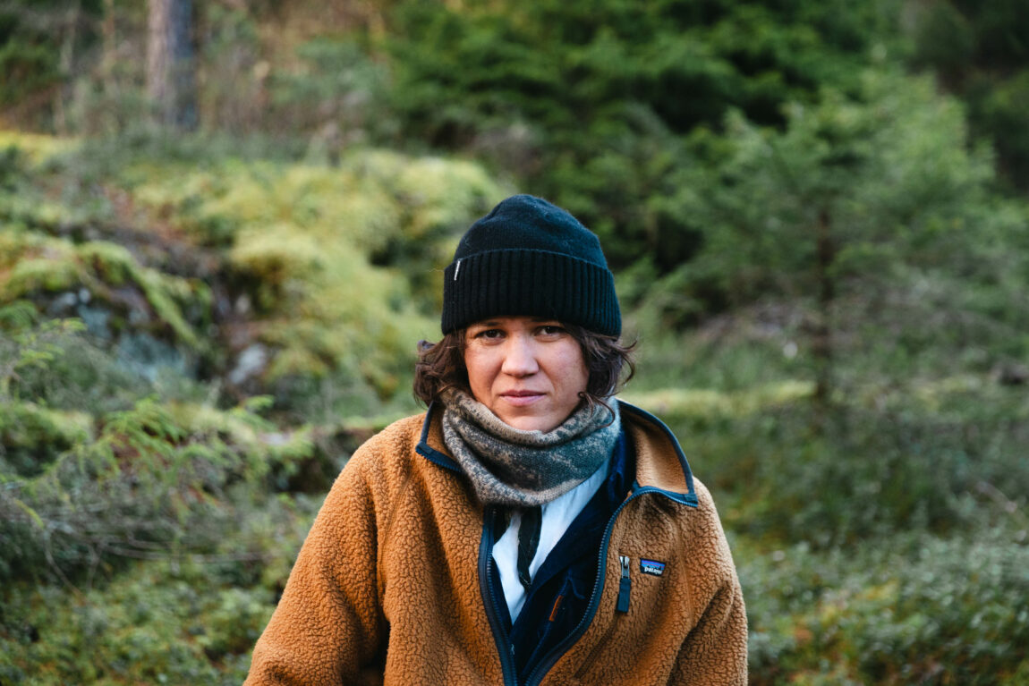 Fay Wildhagen har på seg en oransje jakke-genser og en blå skjorte. Hun har også en grått skjer og en svart lue. Bak henne er det litt grått gress.