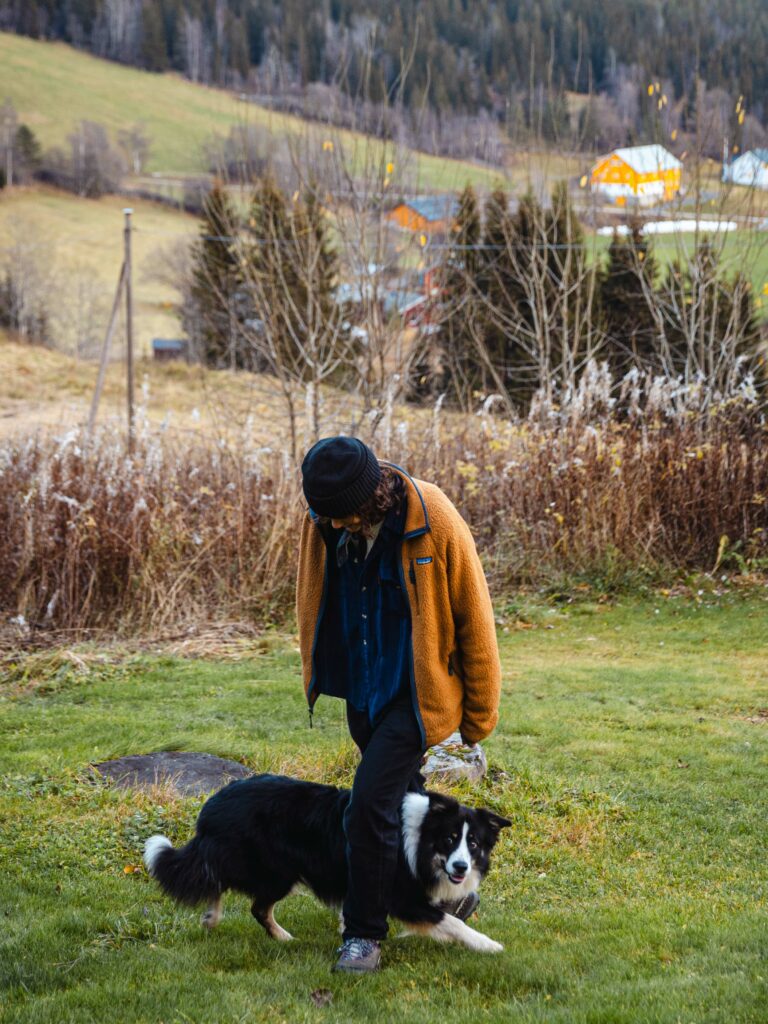 Fay Wildhagen har på seg en oransje jakke-genser og en svart lue. Mellom beina hennes går den svarte og hvite hunden hennes. Den løper i det fri på gresset.