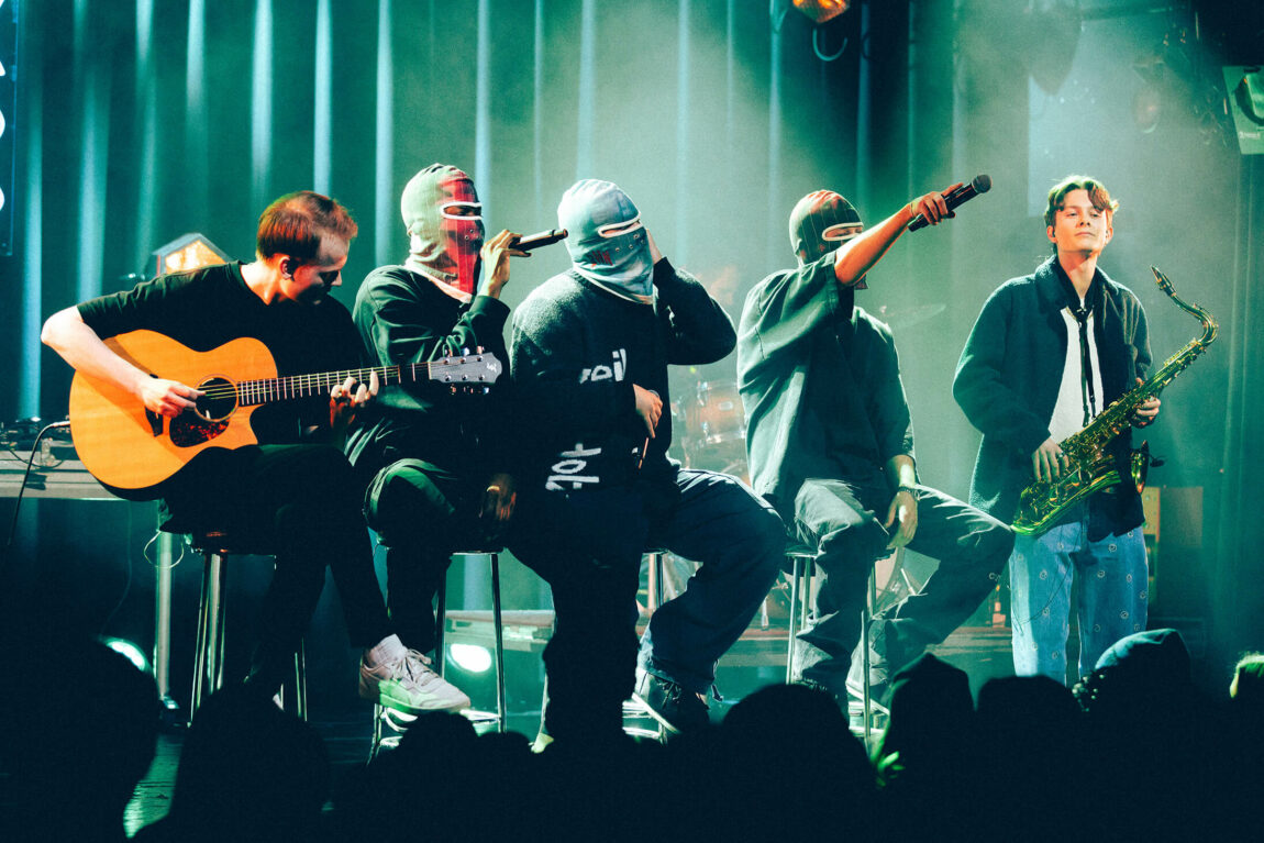 Tre fyrer med masker sitter på krakker med mikrofoner i hendene. På hver sin side sitter en fyr med gitar og en fyr med saksofon.