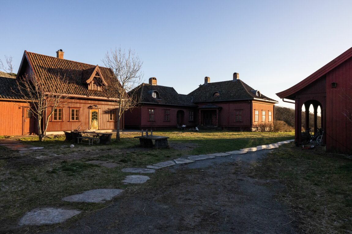 Utsiden av gården. Tre røde hus og en gressplen. Det er skyfri himmel.