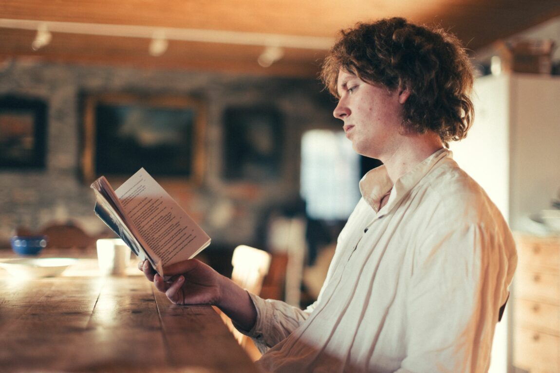 Øde sitter på en stol foran spisebordet i stuen. Han leser en bok. Han har på seg en kremfarget skjorte.