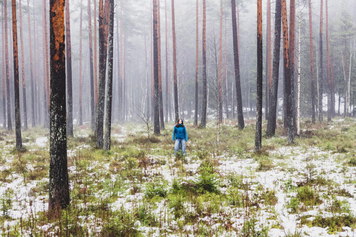 May har på seg en blå jakke og blå bukse. Hun står midt i skogen med masse gress rundt seg. Hun står på avstand, ganske langt unna.