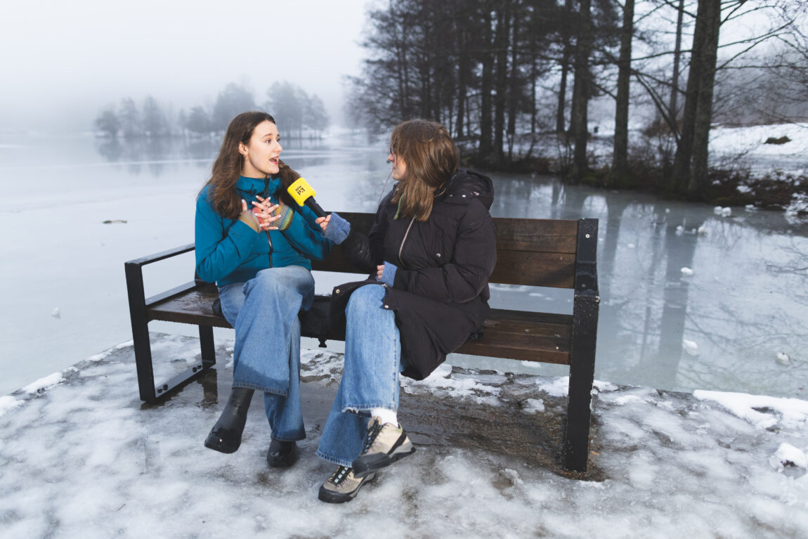 Nina Gjertsen intervjuer May på en benk ved Sognsvann. Det er is og tåke i bakgrunnen. Nina har på seg en svart boblejakke, May har på seg en blå allværsjakke. May ser veldig sjokkert ut.