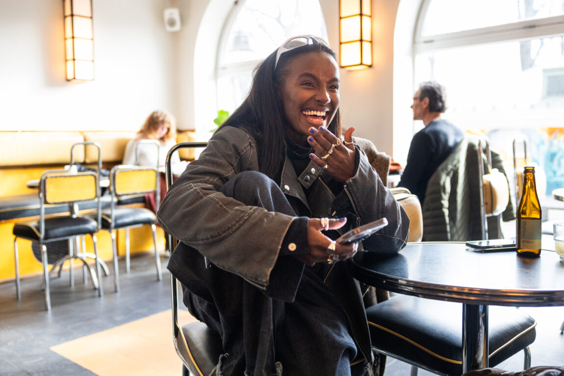 Elia har på seg en svart denimjakke og sølv solbriller på panna. Hun sitter inne på en cafe og smiler og ler.