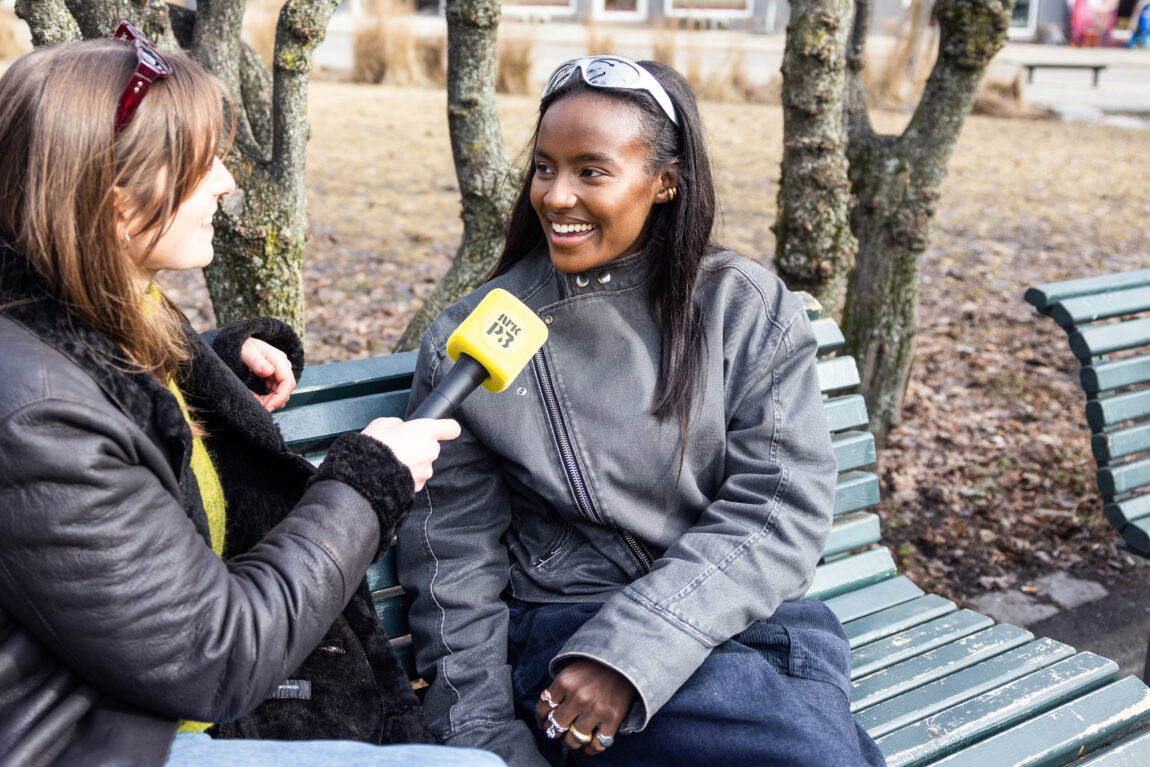 Elia har på seg en svart denimjakke og har hvite solbriller på panna. Hun fniser i vinden. Hun blir intervjuet av Nina Gjertsen.