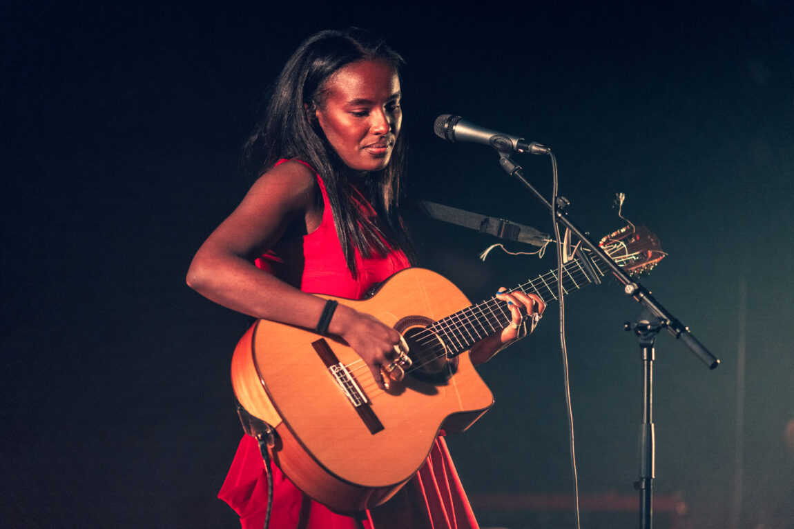 Elia har på seg en rød kjole og spiller gitar på scenen.